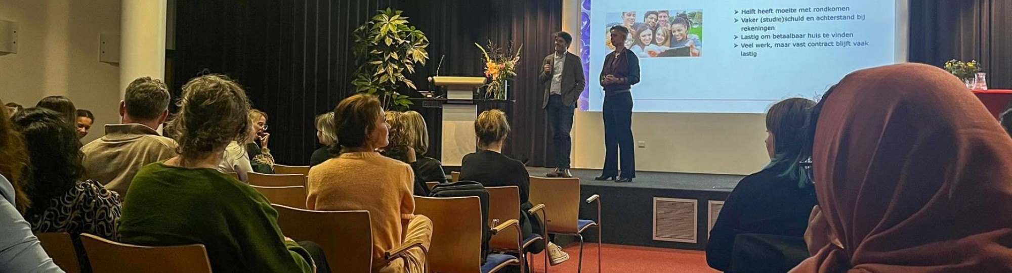 sfeerbeeld van de opening van de week tegen armoede. Mensen zitten op een stoel in een zaal met rode vloerbedekking. In het midden op de foto een podium met daarop Arjan Vliegenthart en Carinda Jansen