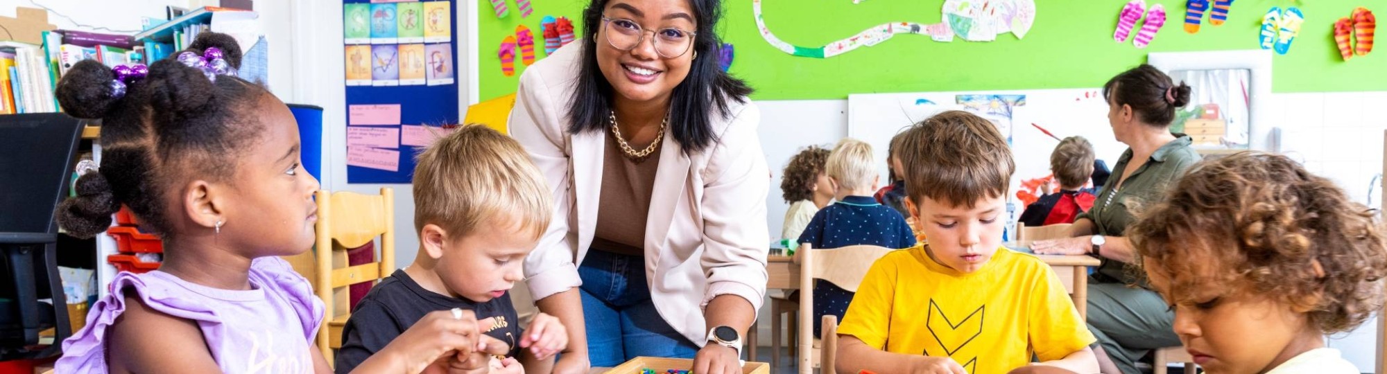 Kinderen leggen een puzzel in de klas en de lerares van de opleiding leraar basisonderwijs helpt hen hierbij