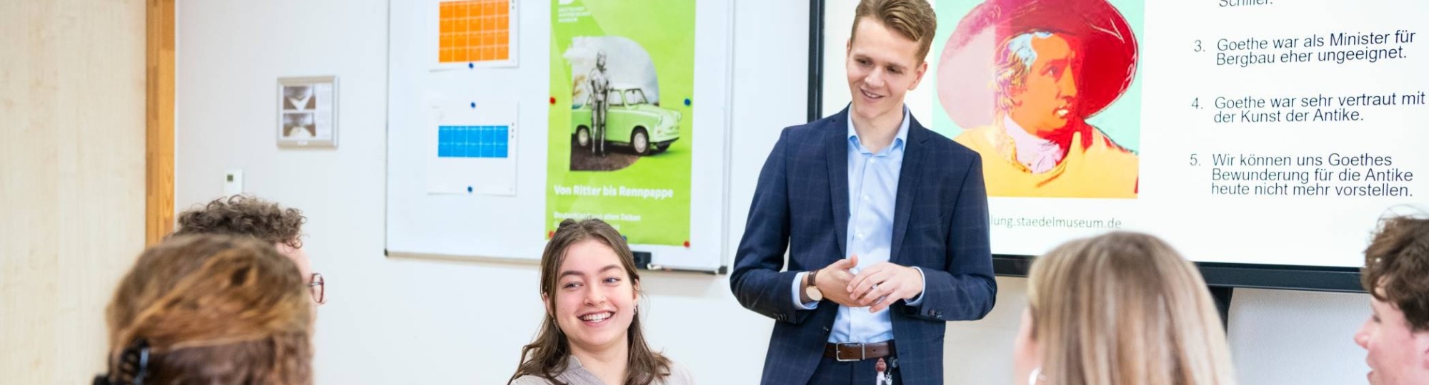 Studenten zitten samen aan tafels met laptops en boeken in het klaslokaal. De leraar staat ervoor en praat met hen mee.
