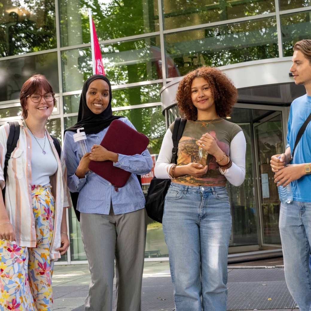 Vier studenten staan bij gebouw LvS10 bij draaideur, buiten. Op een rijtje. 