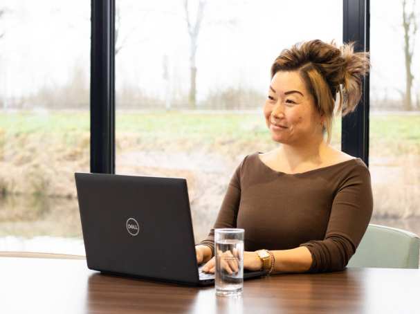 Dame poseert met een glimlach, achter een laptop. Een glas water staat naast haar op het bureau. 