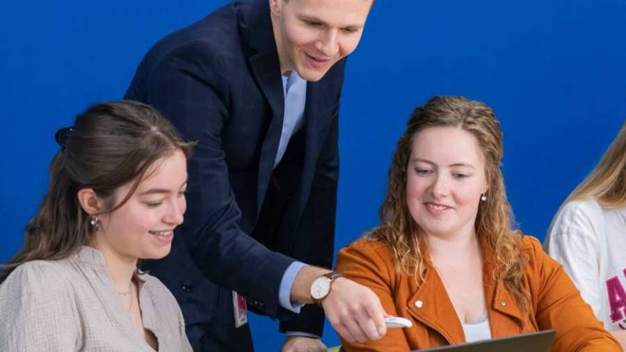 Docent in donkerblauw pak kijkt mee op de laptop bij twee van zijn studenten.
