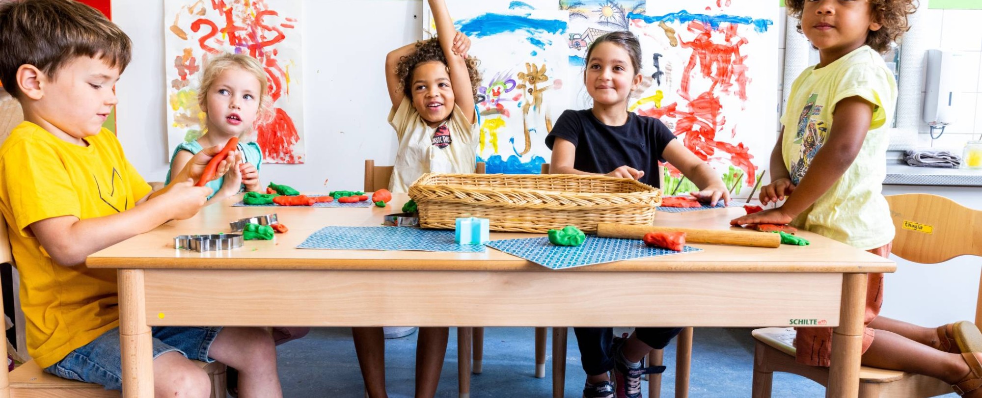 Vijf kinderen zitten aan hun tafel in de klas, waarvan één kind zijn vinger opsteekt om een vraag te stellen. Foto voor de opleiding leraar basisonderwijs