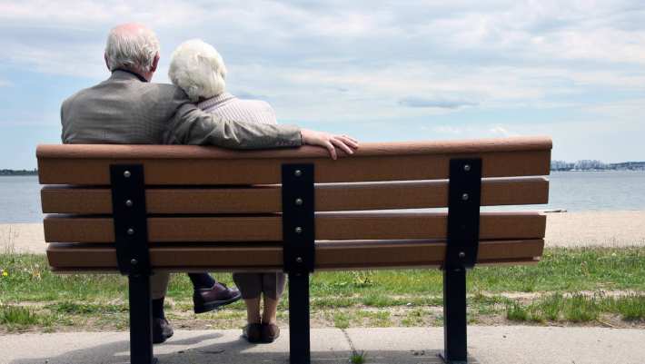 Een ouder echtpaar zit gearmd op een openbaar bankje voor een waterplas, kijkend naar de horizon.    