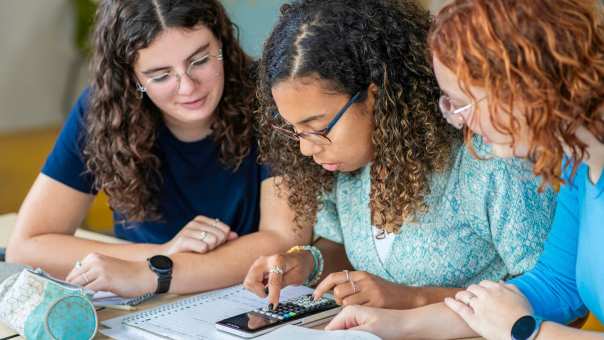 Twee studenten Leraar Wiskunde kijken toe terwijl een andere student een som intypt op de rekenmachine
