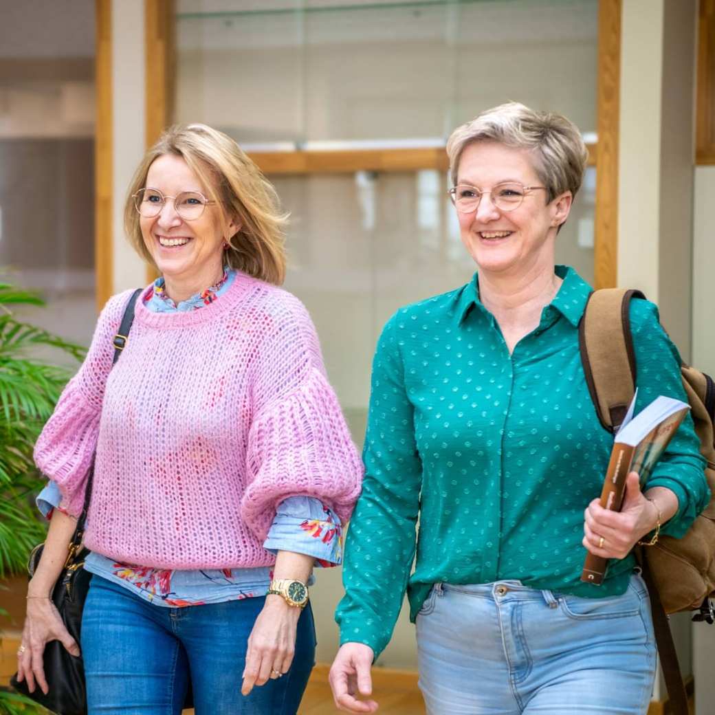 Twee studenten lopen naast elkaar en lachen, terwijl ze door de gang lopen.