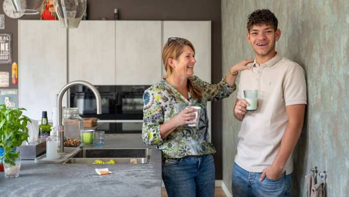 Moeder en zoon lachen en staan in de keuken, terwijl de moeder eeen hand op de schouder van haar zoon legt. 