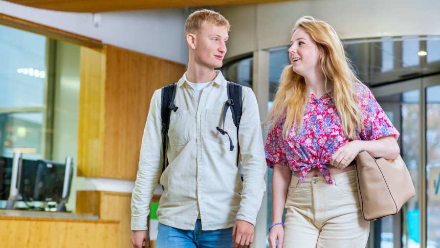 Twee studenten lopen naast elkaar door de gang en vertellen met elkaar.