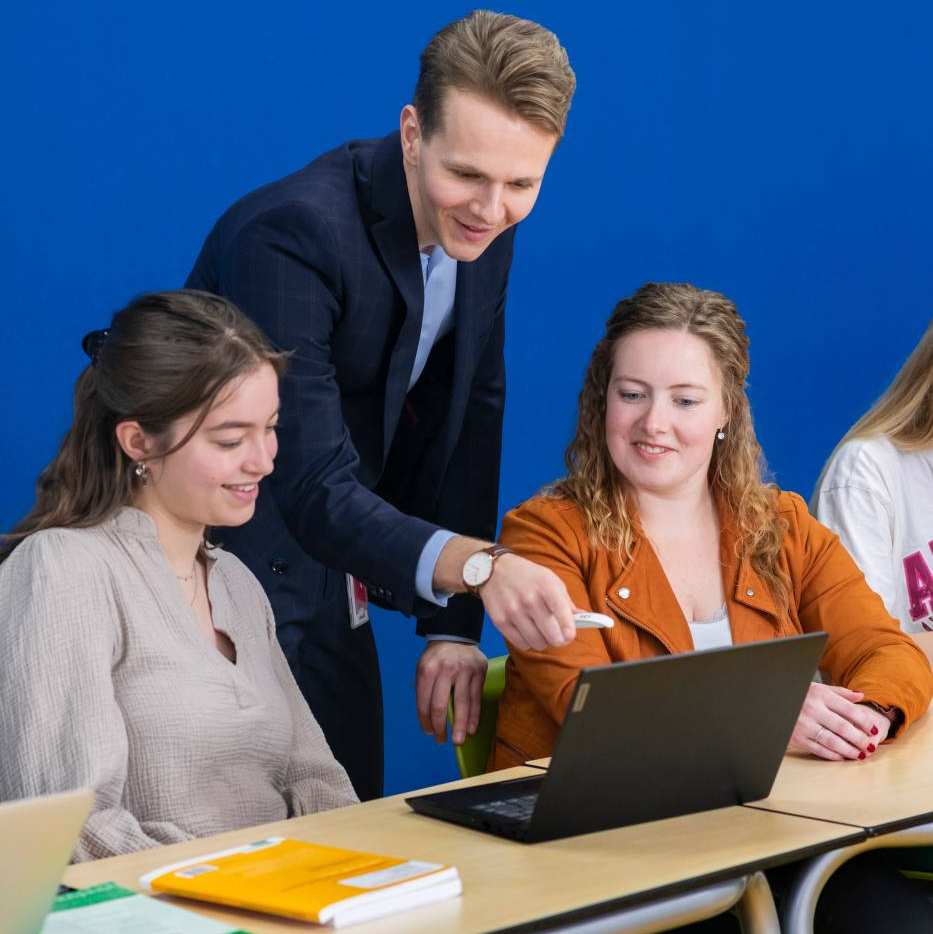 Drie studenten zitten met de docent aan tafel en één van die studenten stelt een vraag aan de docent die aandachtig luistert.