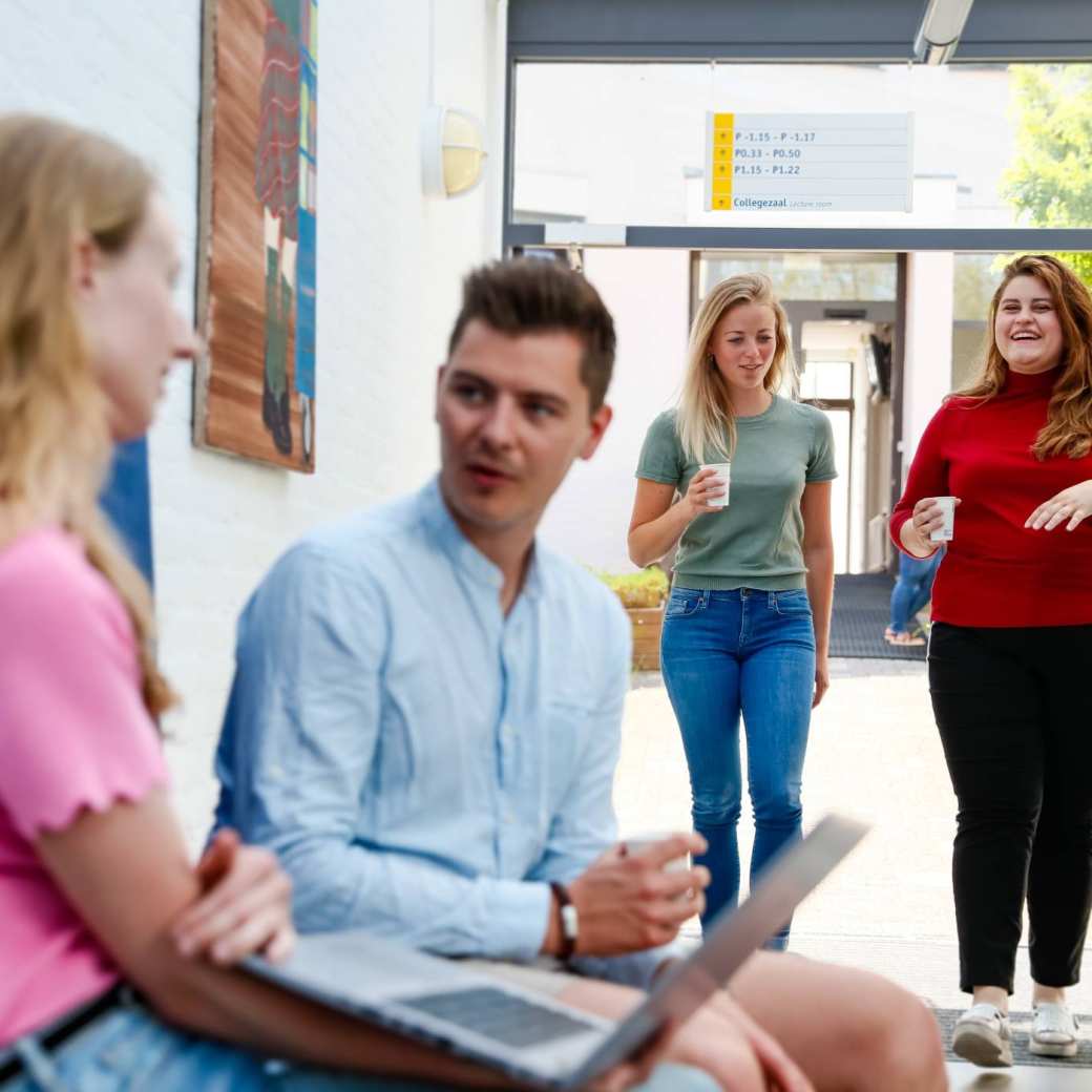 een masterstudent zit op de rand van de tafel met een laptop in z'n hand en legt wat uit aan de rest