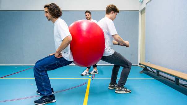 Drie studenten bezig met oefening waarbij twee studenten een roze bal vasthouden met de rug