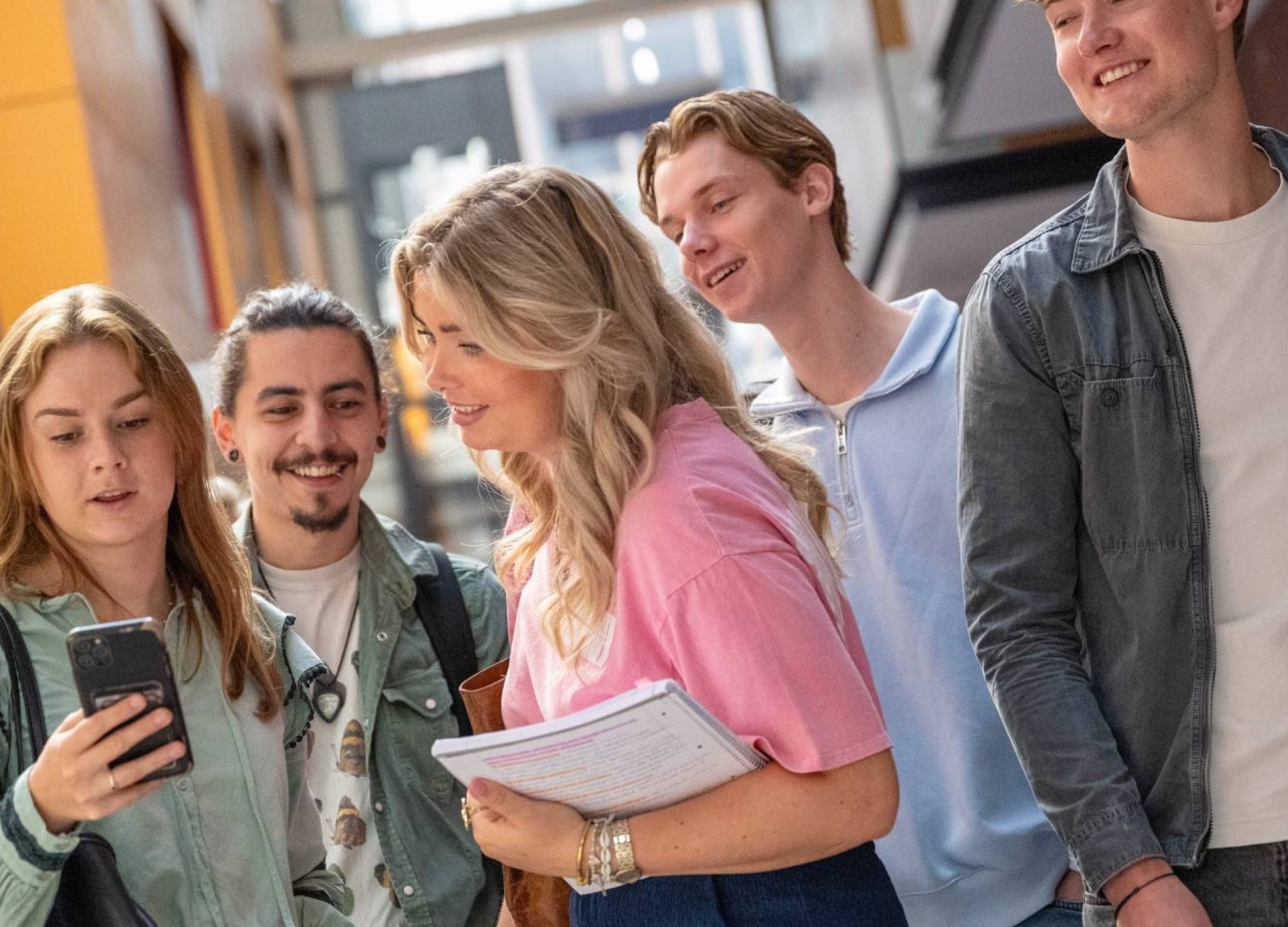 Groepje studenten staan bij elkaar en hebben plezier. Studenten kijkt op haar telefoon, ze staan in de gang. 