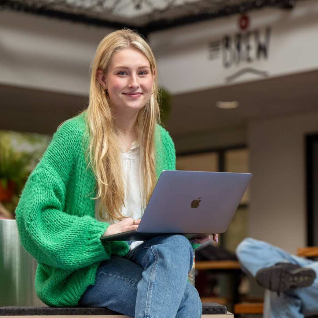 Studente lacht in de camera, heeft laptop op schoot en is aan het werk bij The Brew. 