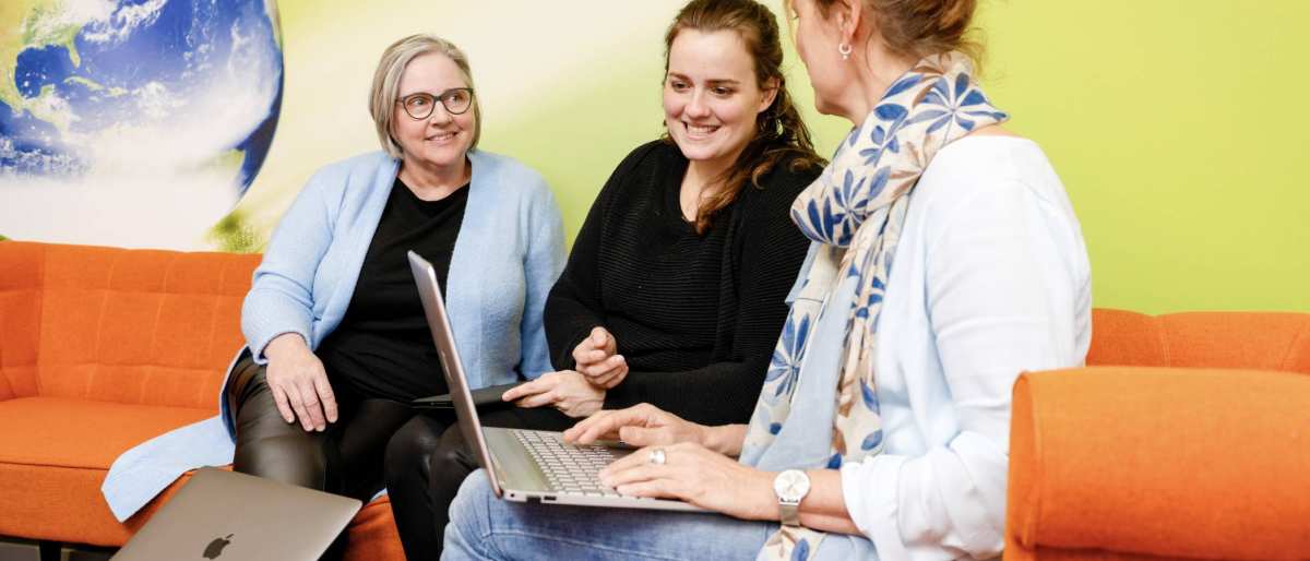 vrouw wijst iets aan op laptop, oranje banken. Wereldbol op achtergrond, tweede laptop op tafel.