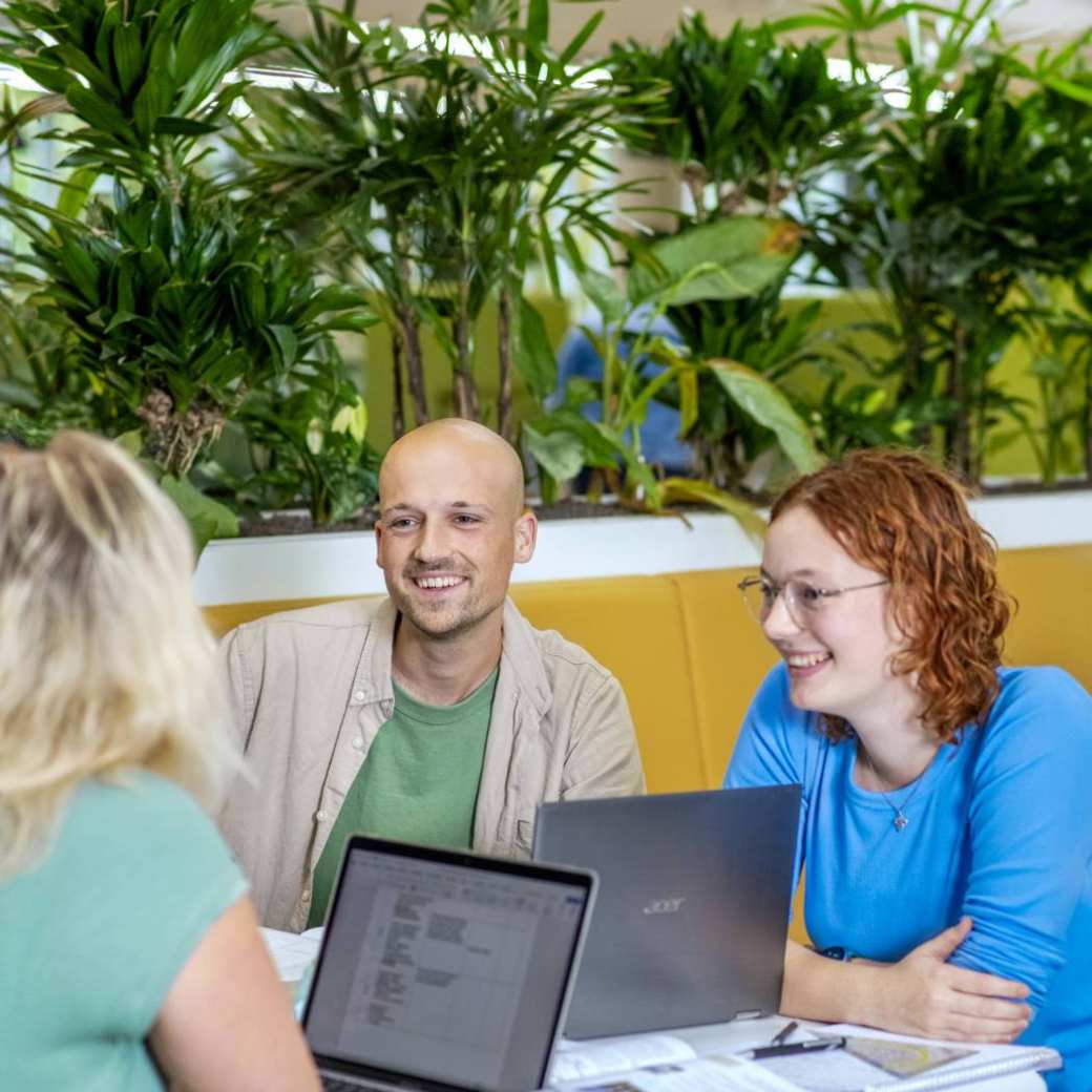 Drie studenten Leraar Wiskunde zitten aan een ronde tafel naar elkaar te lachen.
