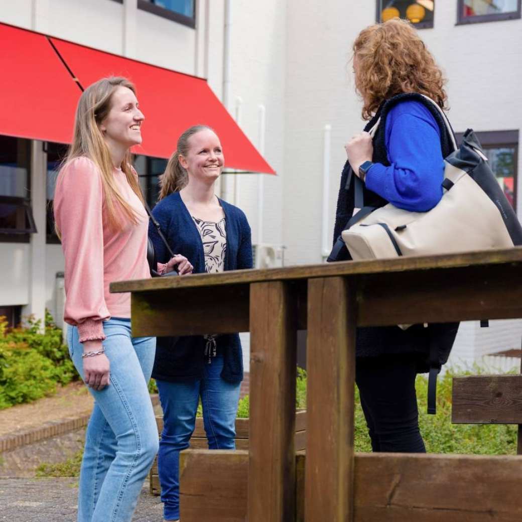 Drie vrouwen in gesprek buiten, achter houten railing.