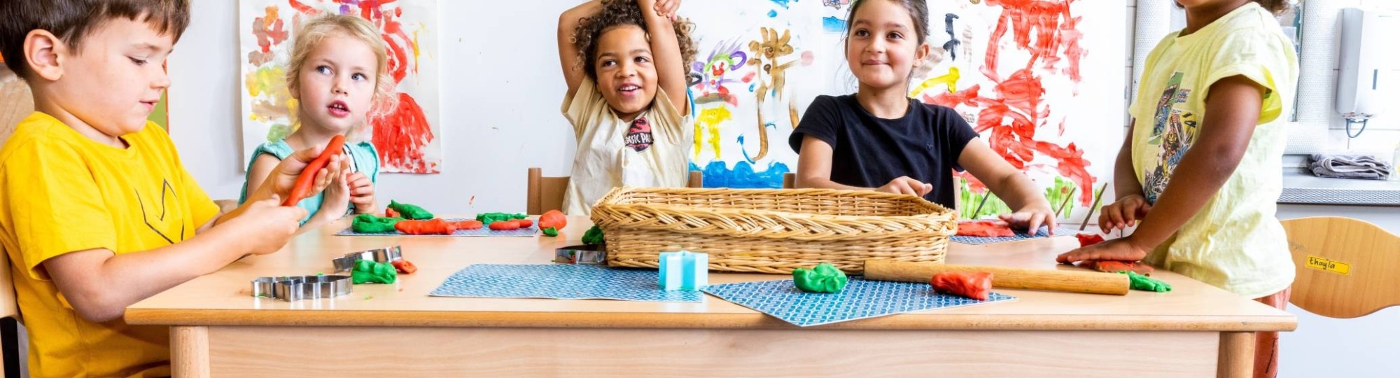 Vijf kinderen zitten aan hun tafel in de klas, waarvan één kind zijn vinger opsteekt om een vraag te stellen. Foto voor de opleiding leraar basisonderwijs