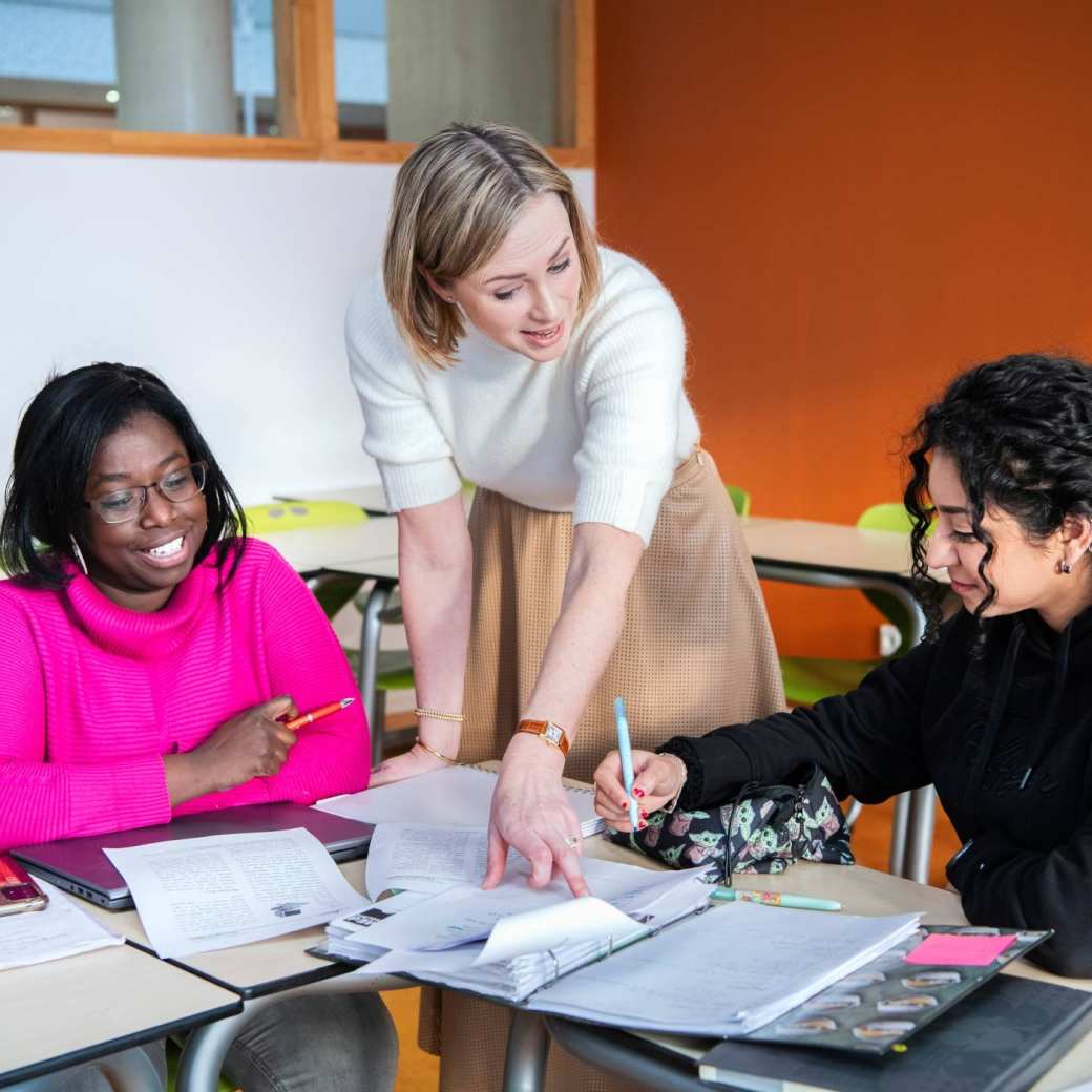 Twee studenten zitten aan tafel. De docente wijst iets aan in het schrift van de studente.