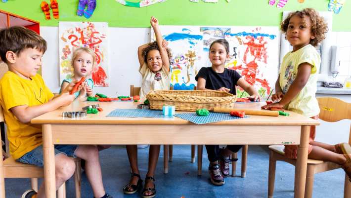 Vijf kinderen zitten aan hun tafel in de klas, waarvan één kind zijn vinger opsteekt om een vraag te stellen. Foto voor de opleiding leraar basisonderwijs