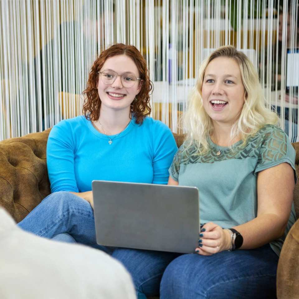 Twee studenten Leraar Wiskunde zitten naast elkaar met de laptop op schoot op de bank. Ze lachen naar de docent die tegenover hen zit.