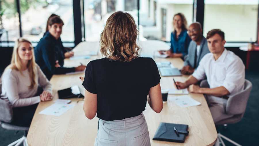 Vrouw, van achteren gezien, leidt een vergadering van een groep collega's.