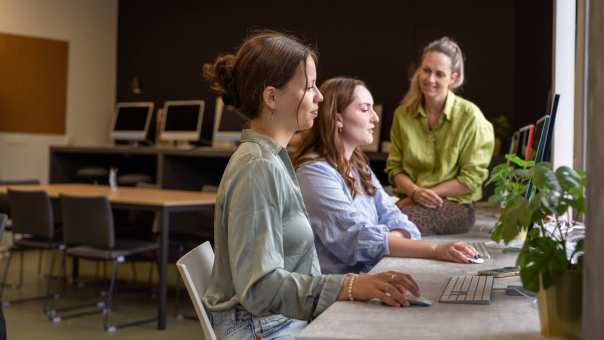 Twee studenten zijn aan het werk op een computer, docent zit er bij. In een klas. 