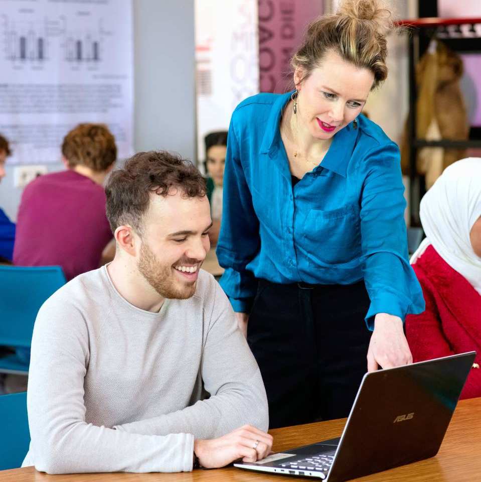 Docent wijst op de computer van een student. Ze lachen naar het scherm.