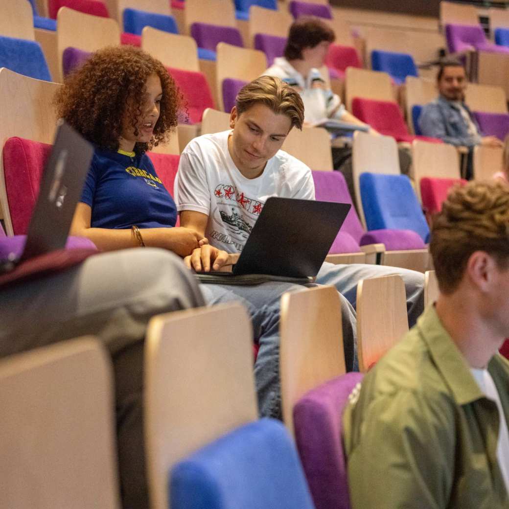 Twee studenten kijken samen op de laptop en overleggen in de collegezaal.