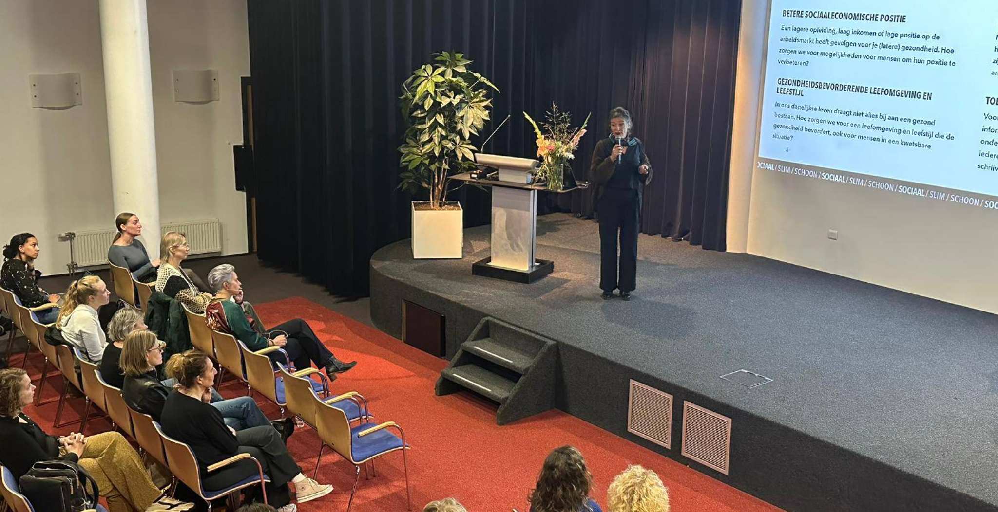 Opening van de week tegen armoede, een zaal gevuld met stoelen en mensen. Op het podium leading lector Lilian Beijer