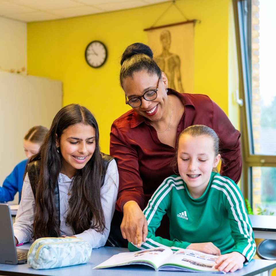 2 lachende leerlingen kijken naar een boek waar de docenten heen wijst op het Citadel College. 