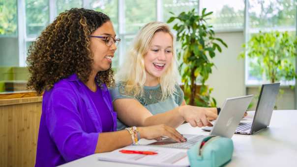 Twee studenten Leraar Wiskunde zitten naast elkaar met een laptop. De ene wijst lachend op het scherm iets aan en de ander kijkt lachend mee.
