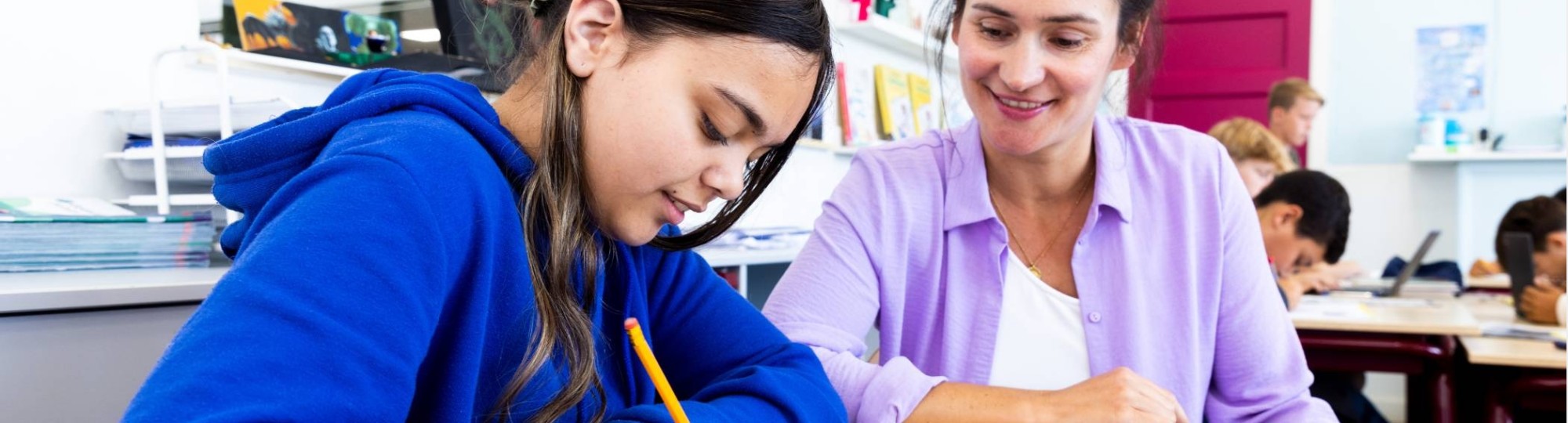 De lerares van de opleiding leraar basisonderwijs geeft persoonlijke uitleg aan leerling aan haar tafel