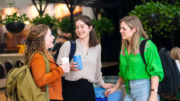 Drie vrouwelijke studenten Leraar Duits kletsen al staand met elkaar, terwijl er twee een drinkbeker in de hand hebben.
