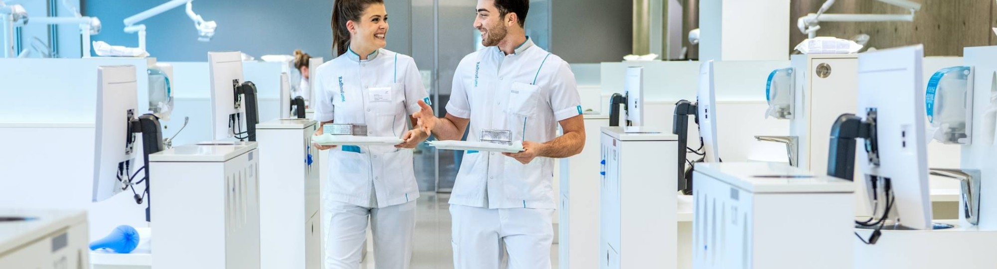 2 studenten kijken elkaar aan en lopen door een ruimte met veel behandelstoelen.