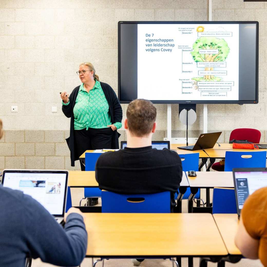 een aantal studenten in een klaslokaal waarbij een docent voor de groep staat