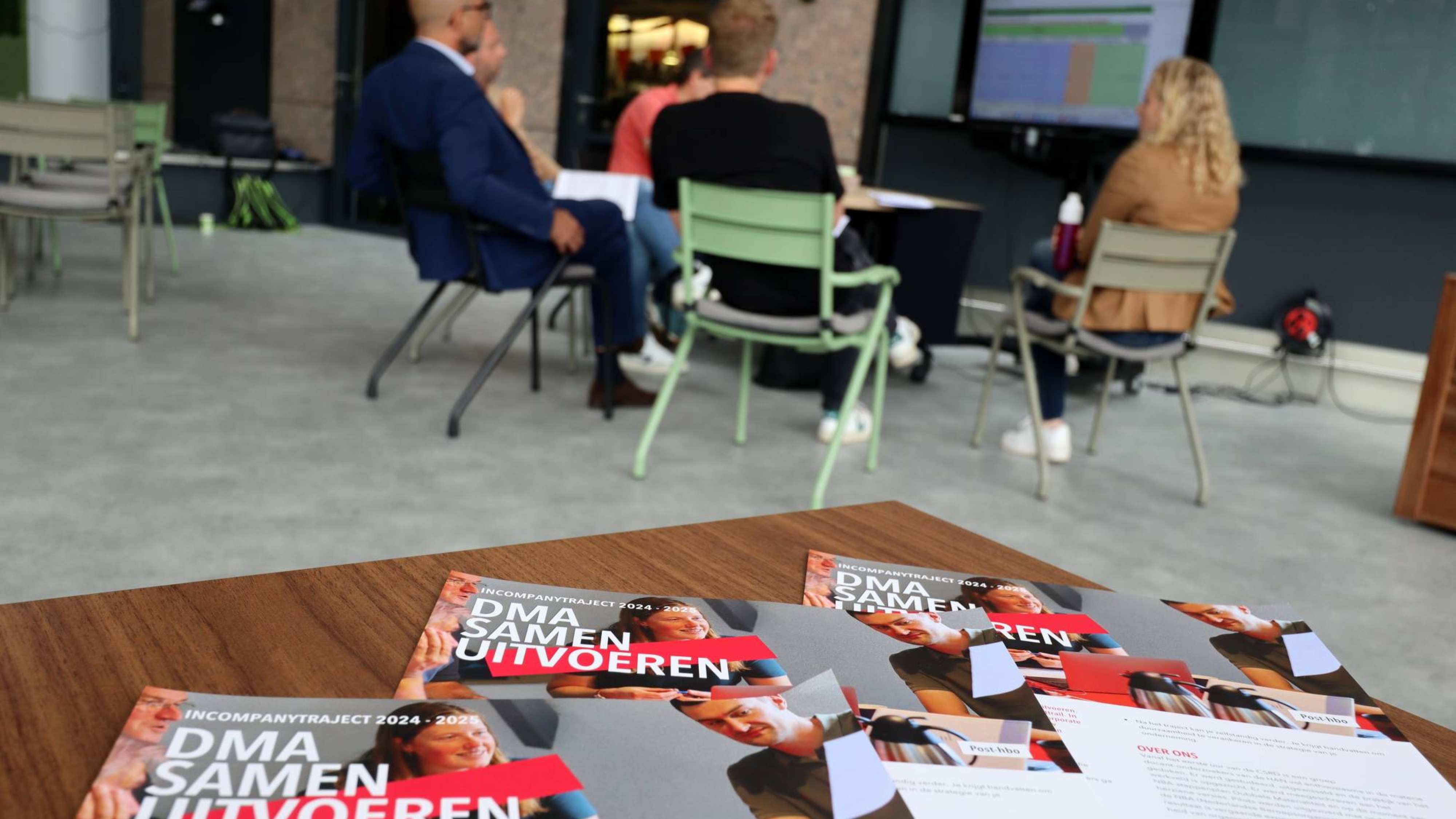 Sessie DMA Samen uitvoeren. Incompany uitgevoerd door de HAN bij Bovemij. Op de foto enkele flyers en een werkgroepje op de achtergrond.