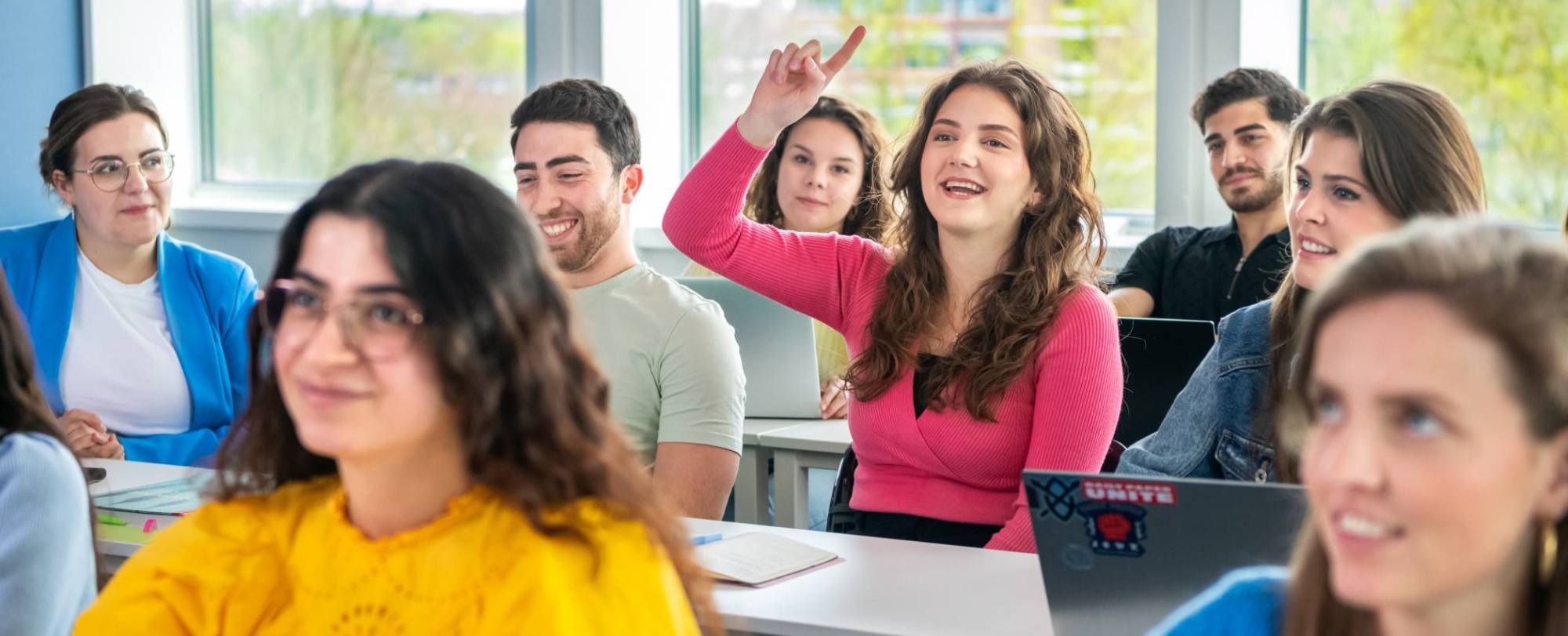 Studenten zitten in de klas, 1 student steekt een vinger omhoog om een vraag te stellen.