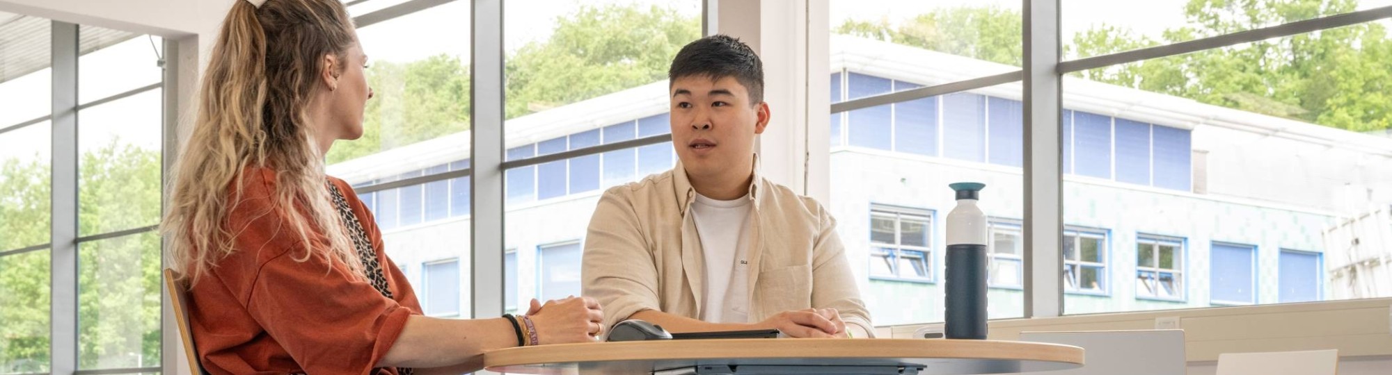 Student in gesprek met docent aan een tafel op podium in hal van gebouw R26, studentbegeleiding. 