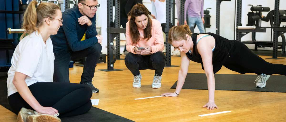 Student staat in een plank positie, 3 studenten kijken toe.