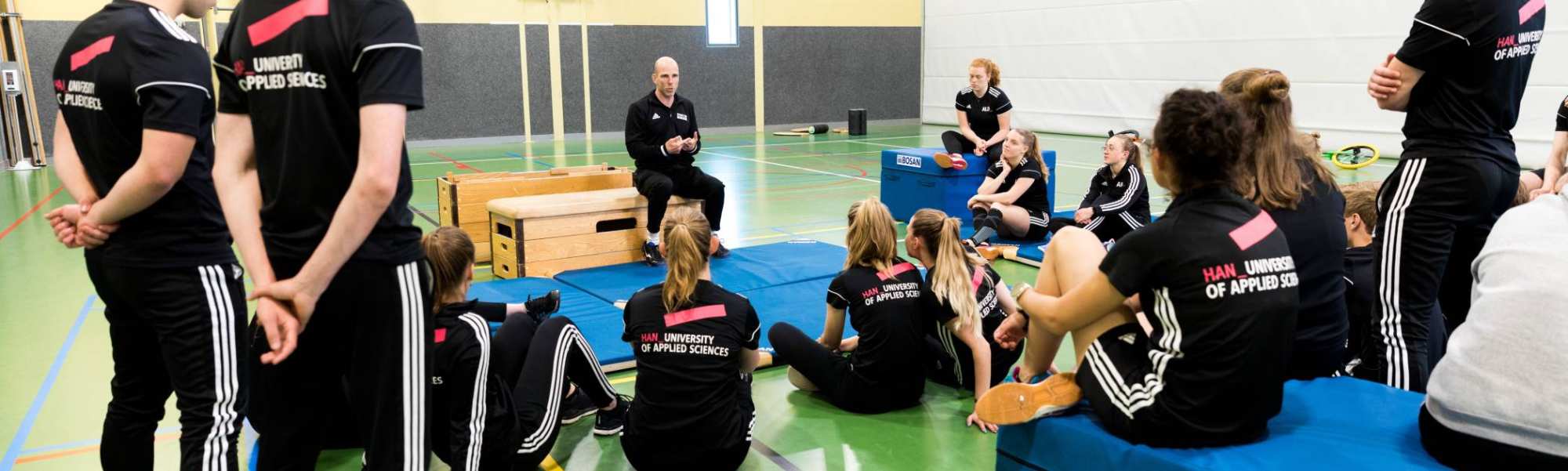 Pabo-ALO studenten zitten in de gymzaal rondom de docent die instructies geeft.