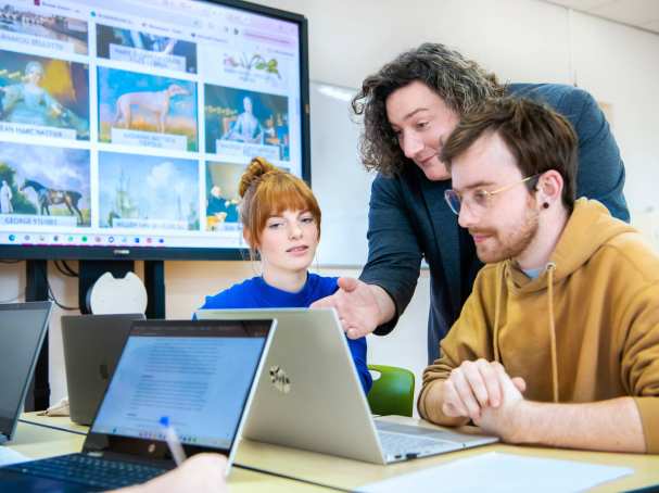 Lerarenopleiding Geschiedenis staat tussen twee studenten in en legt hen iets uit, terwijl iedereen op de laptop voor zich kijkt.