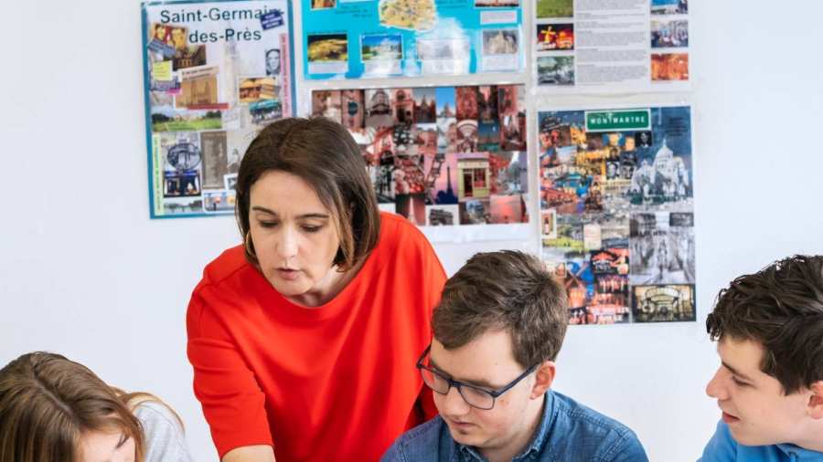 Drie studenten zitten op een rij en de docent staat achter hen. Ze wijst in het boek en de studenten kijken mee.