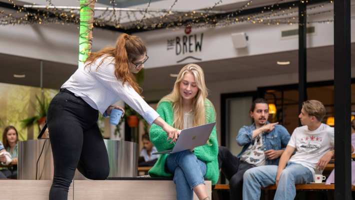 Twee studenten overleggen samen en wijzen naar de laptop, aan het werk bij The Brew.