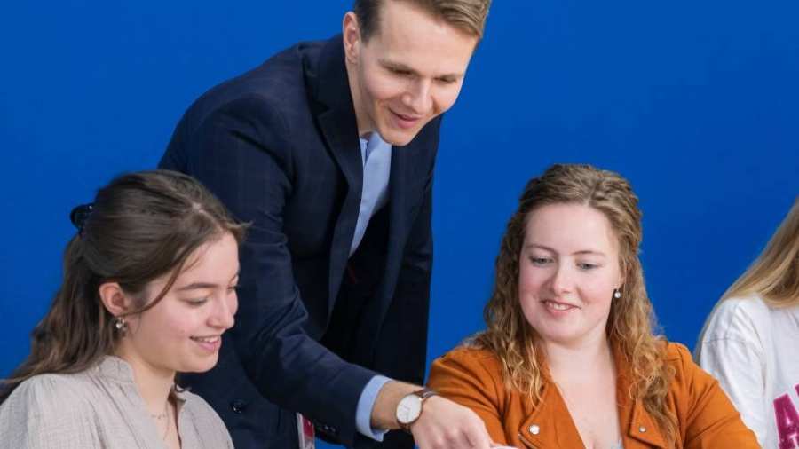 Docent in donkerblauw pak kijkt mee op de laptop bij twee van zijn studenten.
