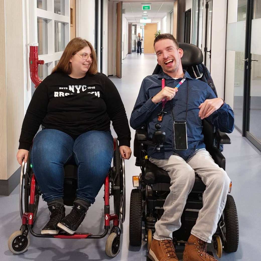 2 mensen in een rolstoel, links een vrouw met donkerblond haar met een bril en rechts een lachende jongen met bruin haar