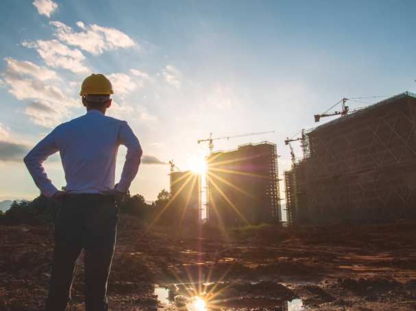 Een man met gele helm kijkt bij een opkomende zon naar gebouwen in aanbouw. Zowel voor bouwkunde als voor civiele techniek bruikbaar.