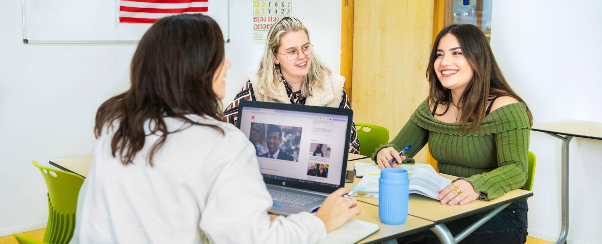 Drie studenten zitten in een Engels lokaal waar de Amerikaanse vlag op de achtergrond hangt