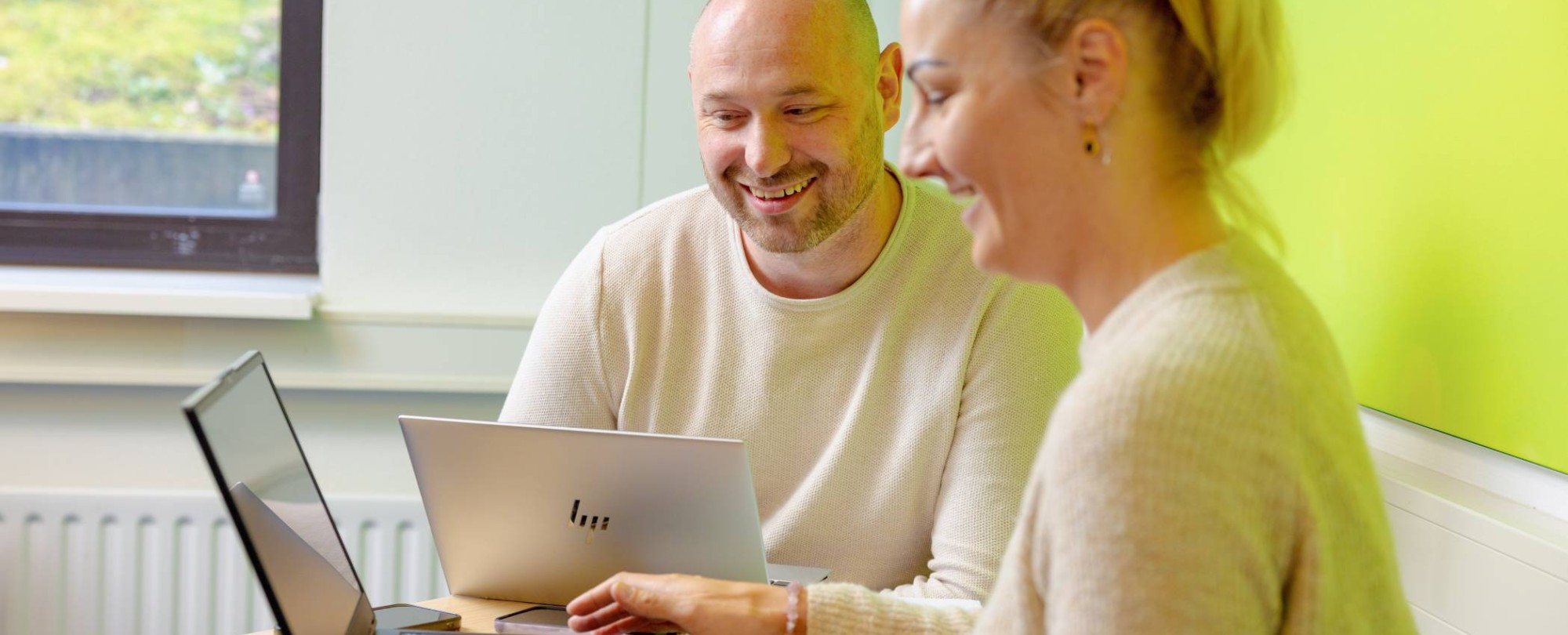 Vrouw en man zitten achter laptop, allebei aan het lachen. Groene muur op achtergrond.