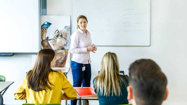 Student leraar Duits in oranje/bruin jasje geeft een lesje aan haar medestudenten. Ze staat voor een PowerPoint.
