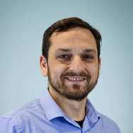 Portrait headshot. The picture is taken in front of a blue background and the subject is wearing a blue collared shirt. His hair is dark, he has a beard and brown eyes. He is facing the camera, with his shoulders slightly towards the right side of the photo.